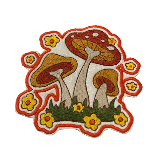 The image shows a handmade embroidered iron-on patch featuring three red and yellow mushrooms with white spots, surrounded by small yellow flowers with orange outlines. The mushrooms are depicted with green grass at their base, and the patch has a white background with an orange border. The design is colorful and detailed, giving it a vibrant and whimsical appearance.
