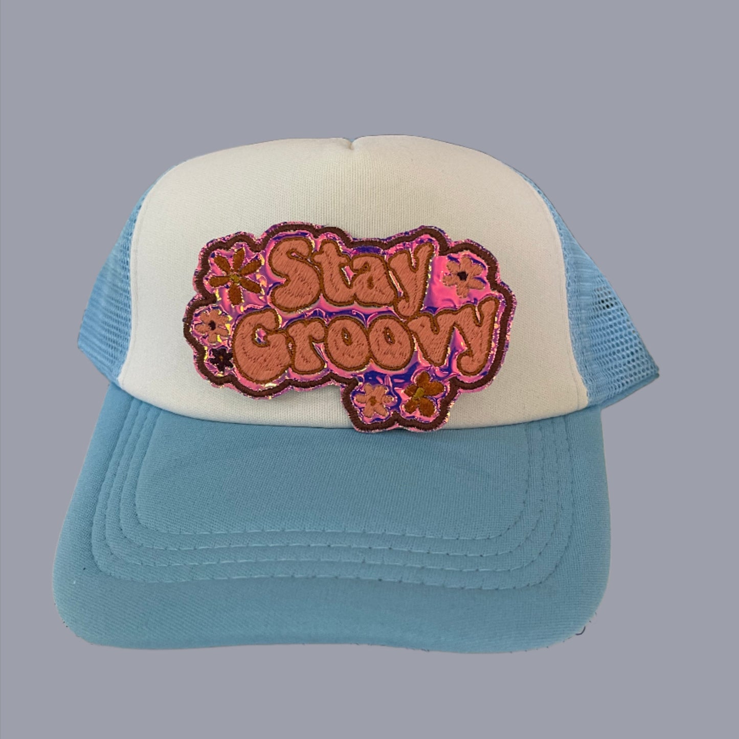 Iron-on patch featuring "Stay Groovy" lettering in a retro font, set against a holographic pink background with playful flowers in shades of peach, pink, and purple, outlined in dark pink, and a glittery border.