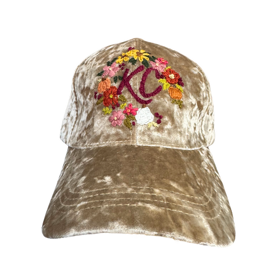 Hand-embroidered floral wreath around KC logo on beige velvet hat.

