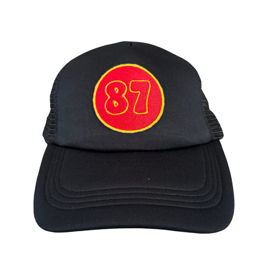 Front view of a black trucker hat featuring an embroidered red and yellow "87" patch, representing Kansas City Chiefs' Travis Kelce.

