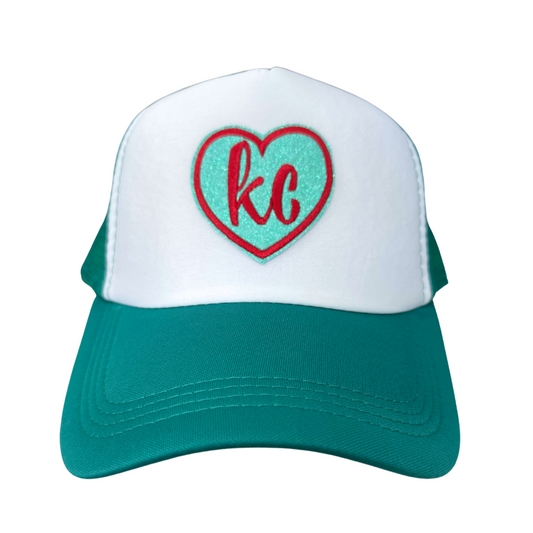 Front view of a teal and white trucker hat featuring a heart-shaped "KC" embroidered patch in red and teal, inspired by KC Current team colors.

