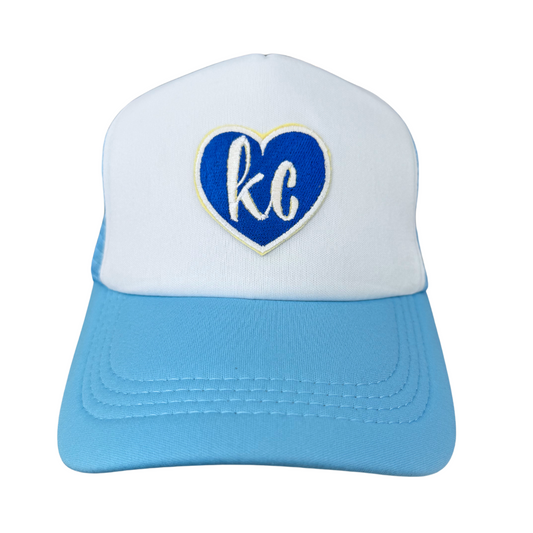 Front view of a trucker hat with a heart-shaped "KC" embroidered patch in Kansas City Royals team colors on a white and blue hat.

