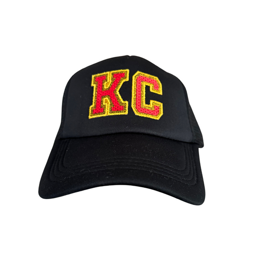 Front view of a black adult-sized trucker hat featuring an embroidered "KC" patch in red and yellow, inspired by Kansas City Chiefs colors.

