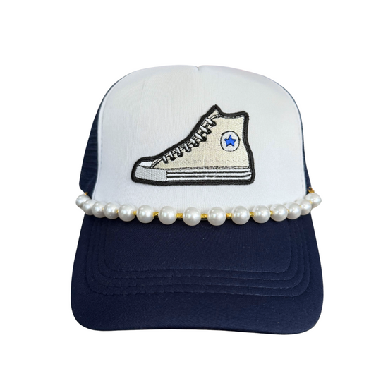 Front view of a navy and white trucker hat with an embroidered Chuck Taylor sneaker patch and pearl embellishments, symbolizing support for Kamala Harris’s presidential run.

