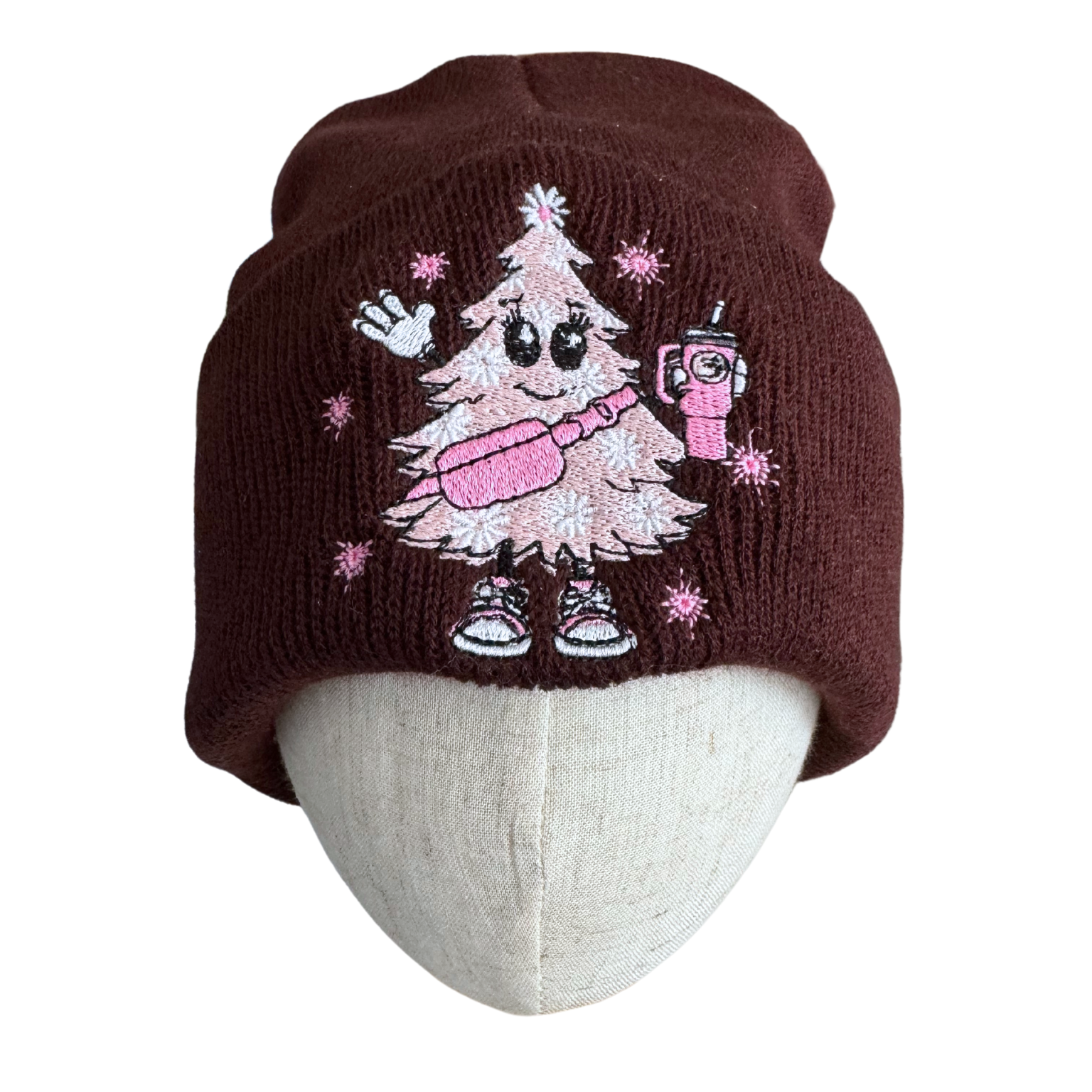 Brown beanie with a pink Christmas tree embroidery wearing sneakers and holding a festive drink.

