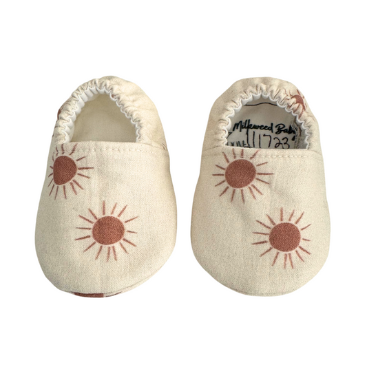 Baby moccasins with a neutral beige base featuring rust-colored sunburst prints, handmade with vegan materials and an elastic back for a snug fit, size 0-3 months, ready to ship.

