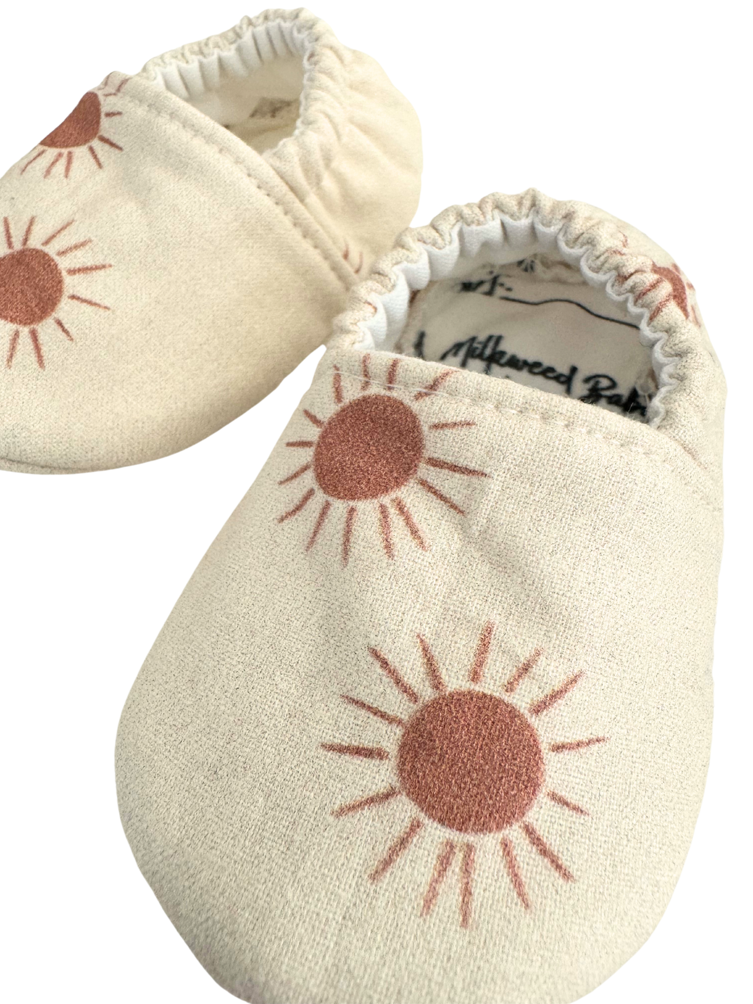 Baby moccasins with a neutral beige base featuring rust-colored sunburst prints, handmade with vegan materials and an elastic back for a snug fit, size 0-3 months, ready to ship.

