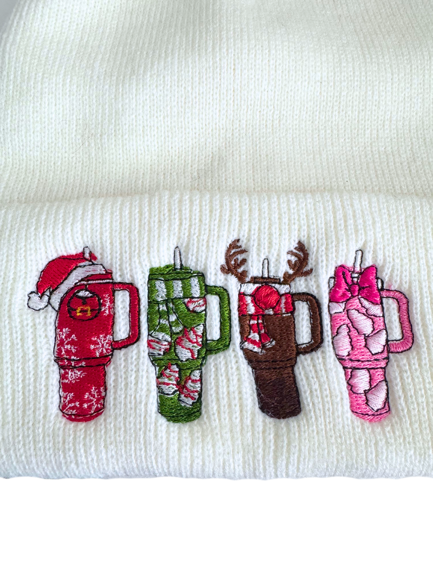 White beanie with embroidered holiday-themed Stanley-style cups, including designs of Santa, reindeer, candy canes, and marshmallows.
