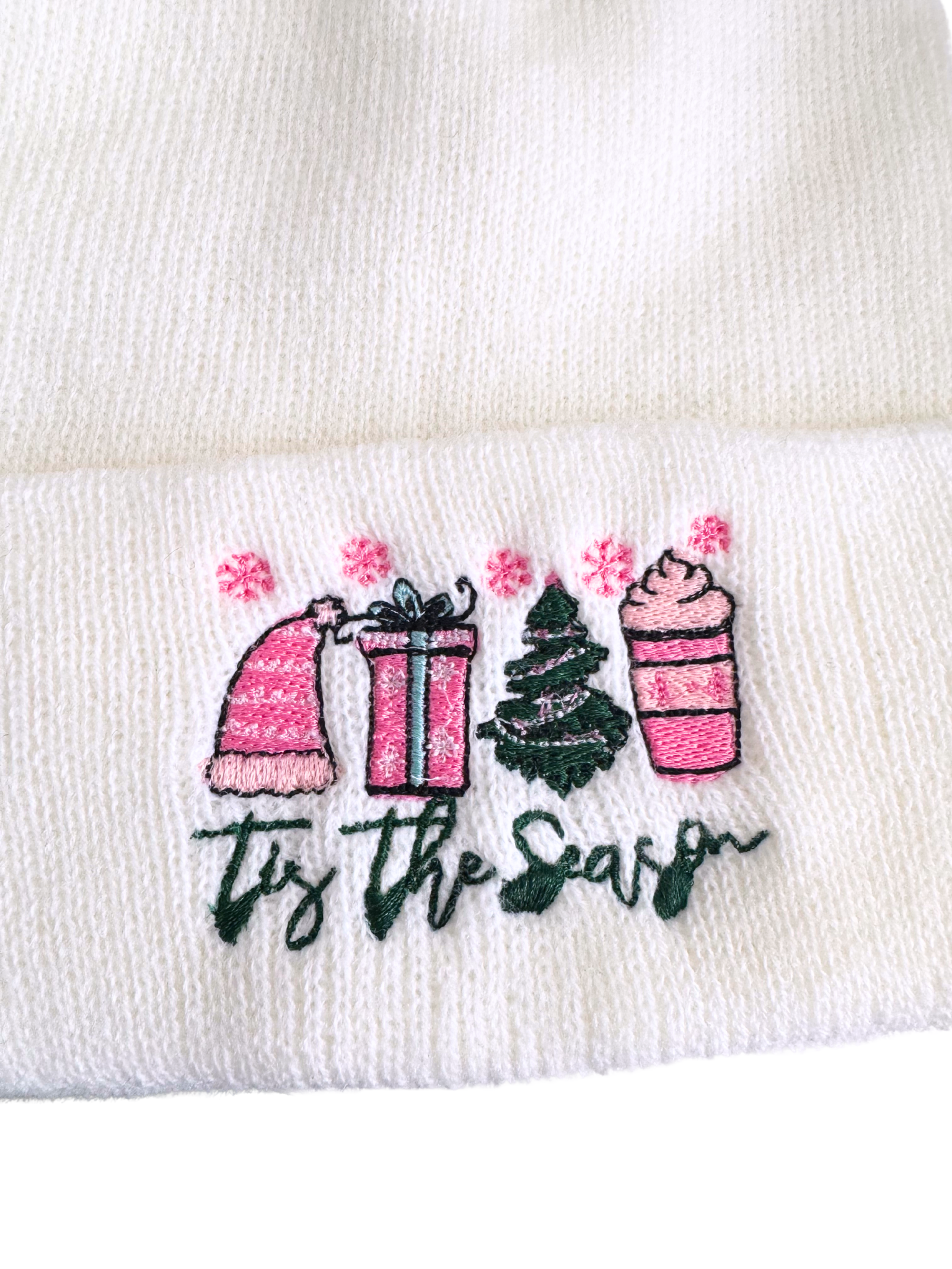 White beanie with "Tis The Season" festive embroidery featuring a pink hat, gift box, Christmas tree, and hot cocoa cup.

