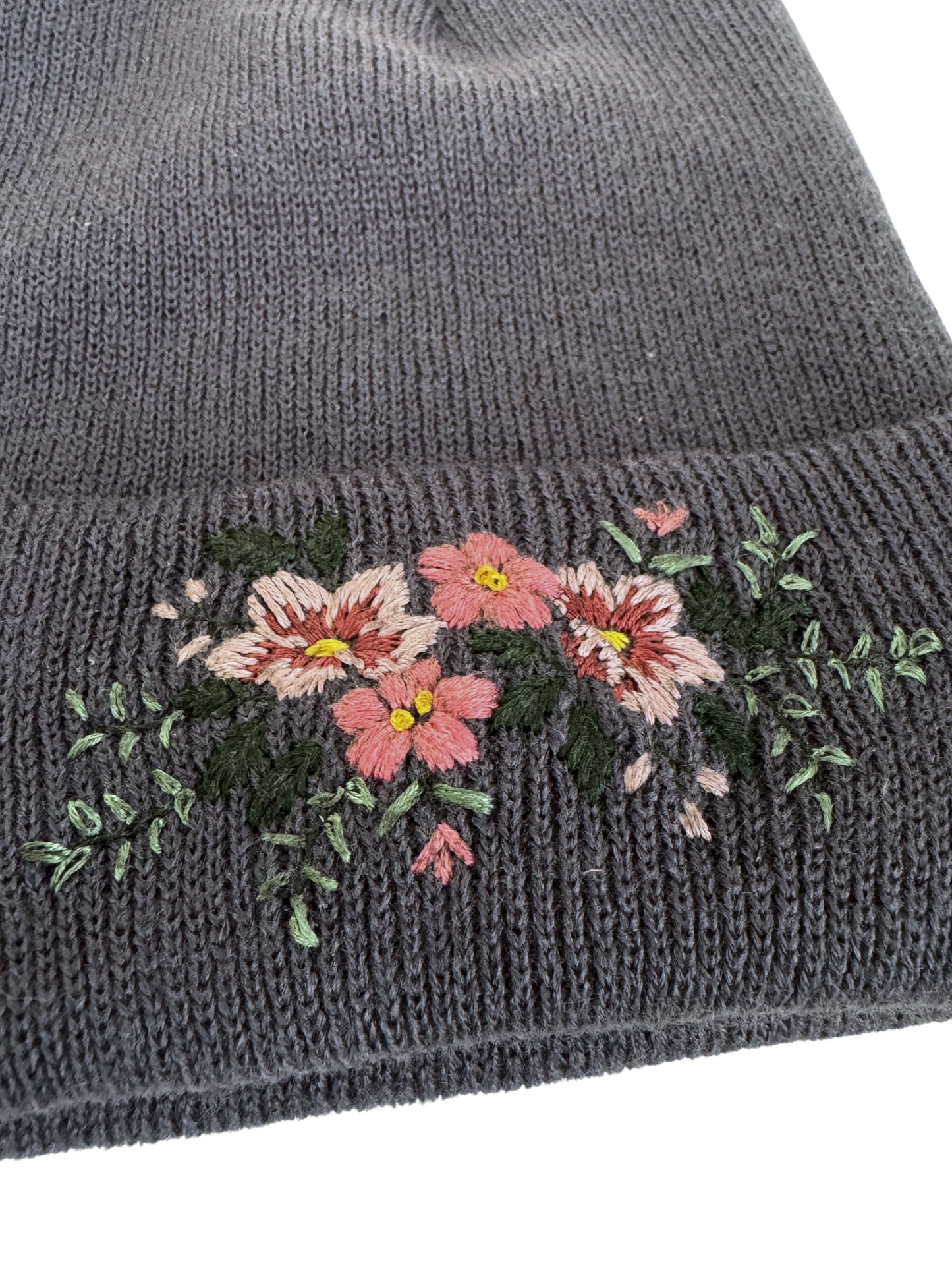 Close-up of hand-embroidered floral design on a blue/gray knit beanie, showcasing intricate craftsmanship.
