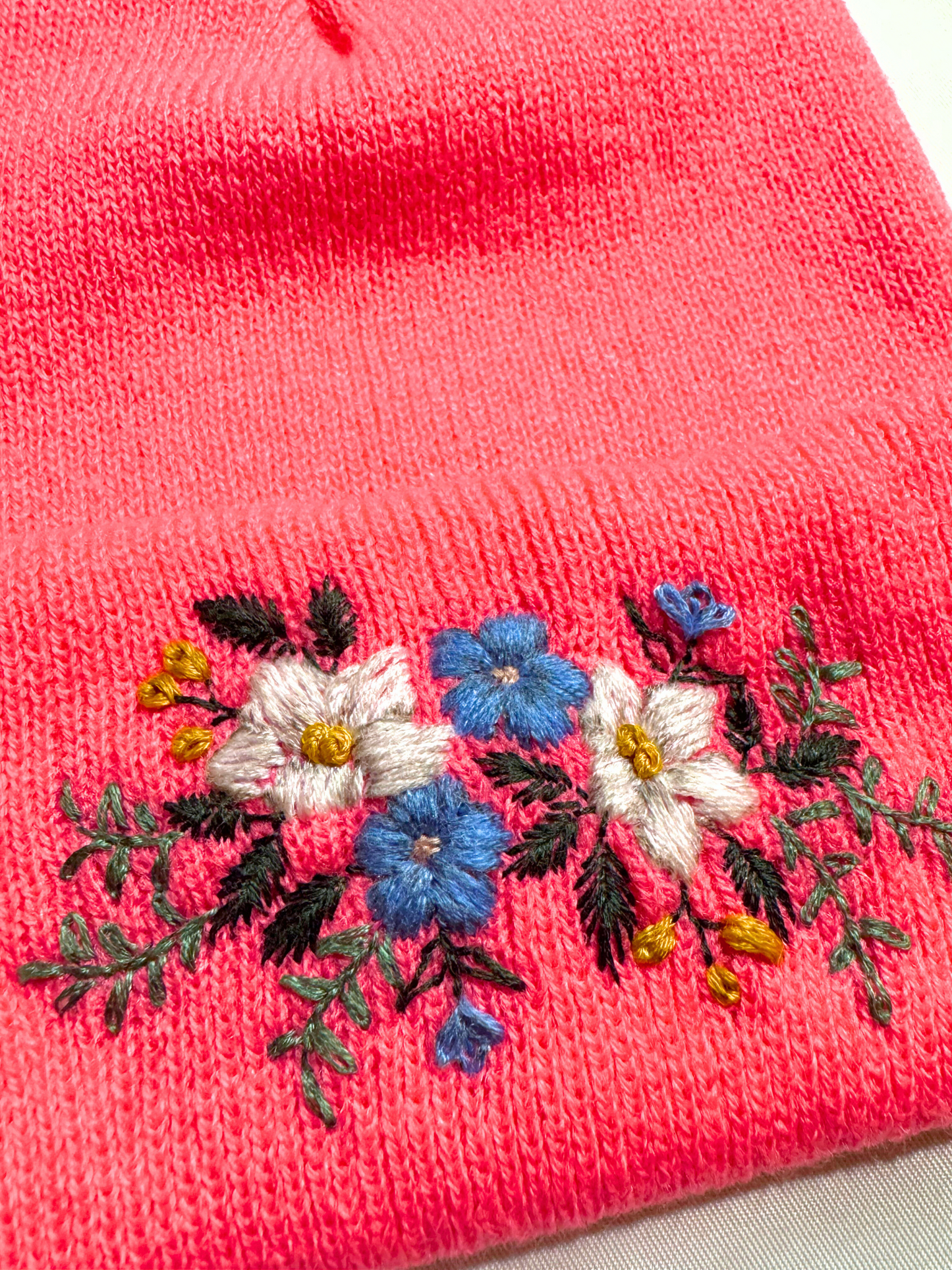 Close-up of embroidered blue and white flowers with greenery on a hot pink knit beanie, highlighting intricate stitching.
