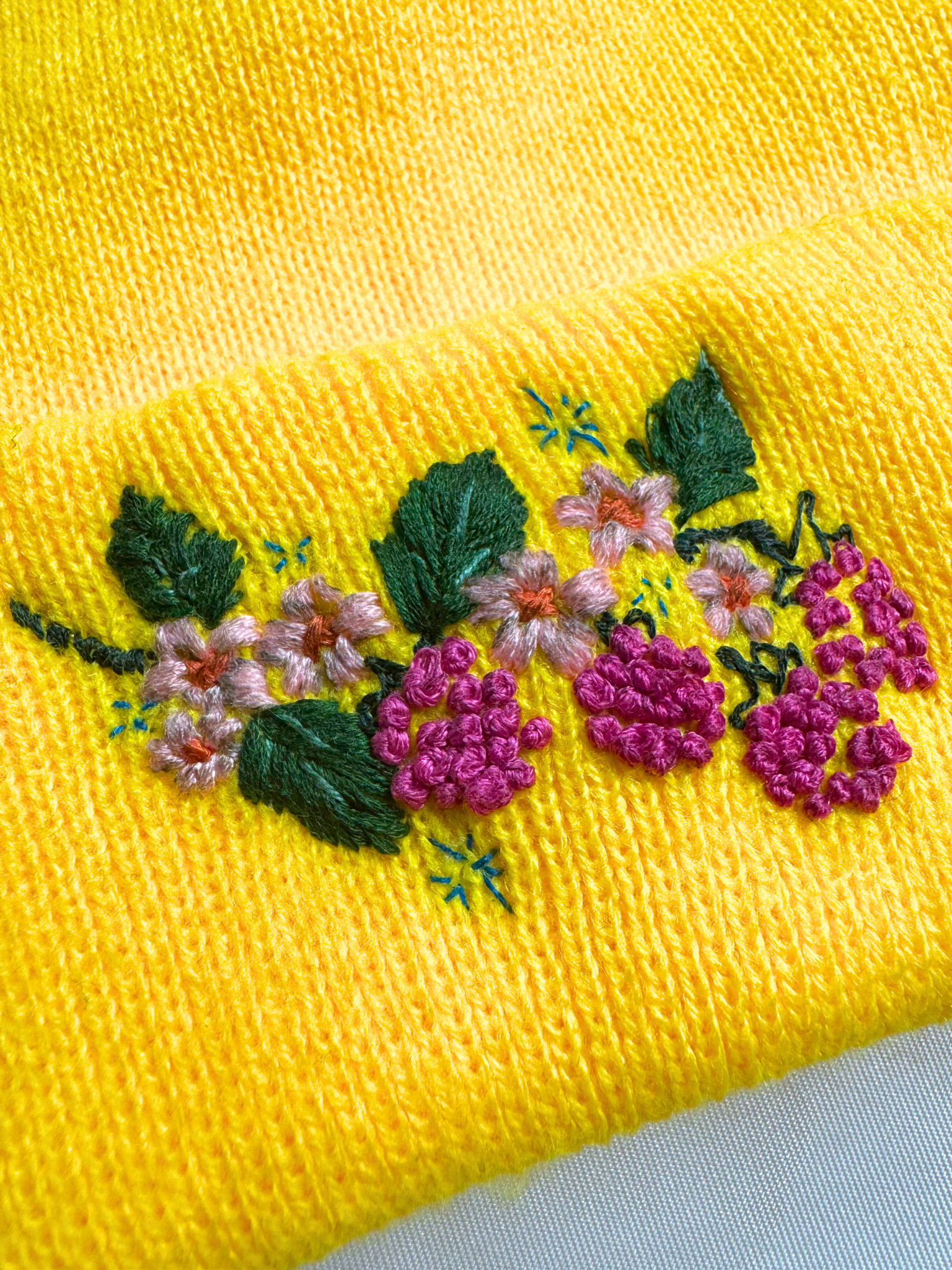 Close-up of embroidered berry and floral design on a yellow knit beanie, highlighting vibrant colors and intricate stitching.
