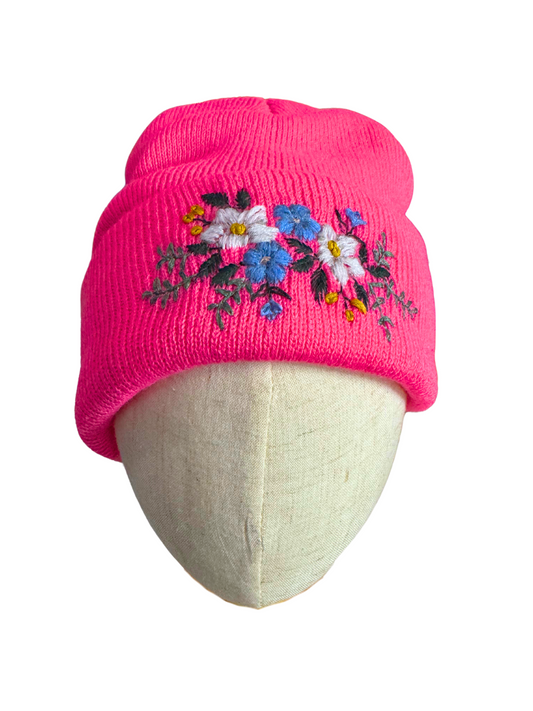 Bright hot pink knit beanie with detailed hand-embroidered floral design in blue, white, and yellow.
