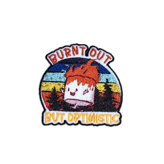 "Burnt Out But Optimistic embroidered patch featuring a happy burnt marshmallow on a retro sunset and forest background."
