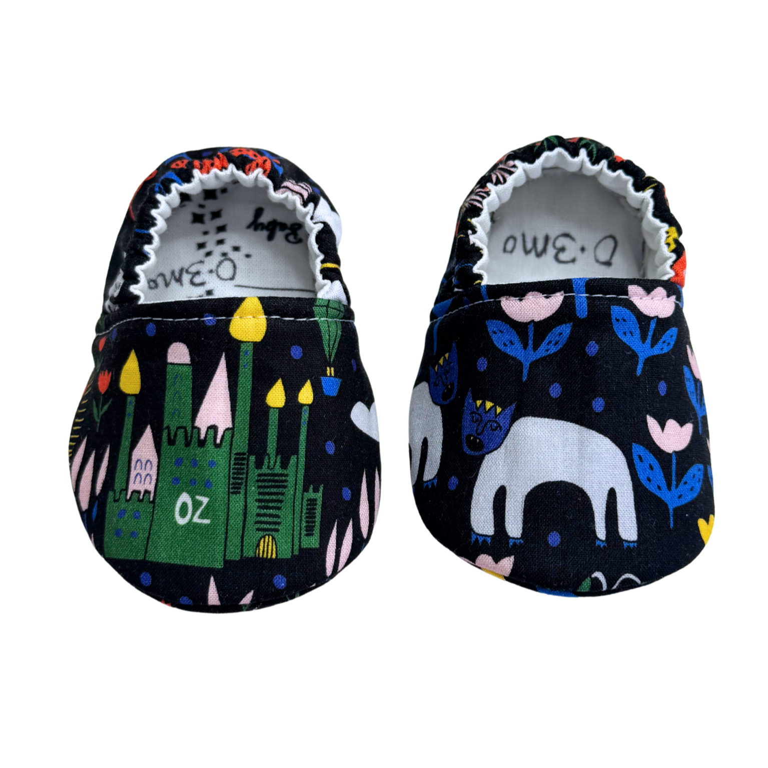 Handmade baby moccasins featuring an Emerald City and Wizard of Oz-inspired design on a navy background, with castles and enchanted creatures, size 0-3 months.