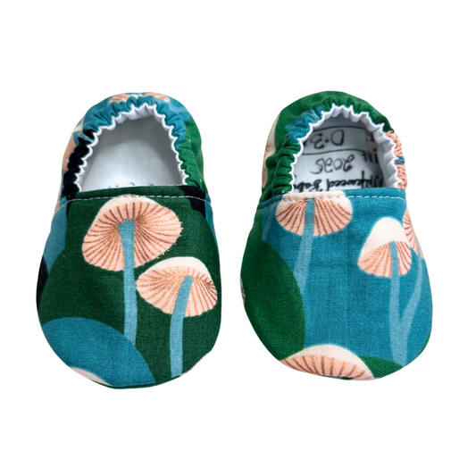 Handmade baby moccasins with a forest-inspired mushroom print in shades of green, blue, and peach, featuring an elastic back and soft soles, size 0-3 months.