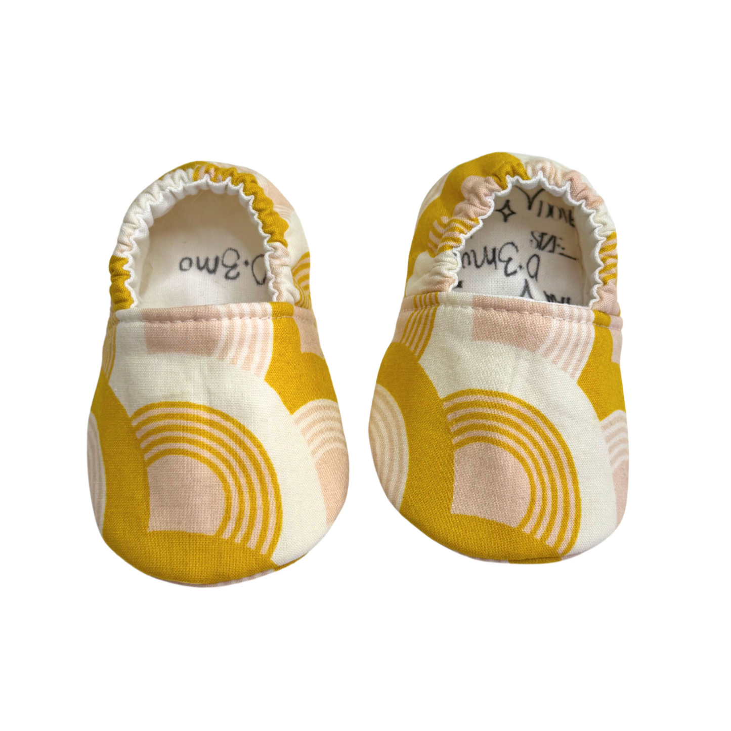 Handmade baby moccasins featuring a mustard yellow and blush pink rainbow print, soft sole, and elastic back for a secure fit, size 0-3 months.