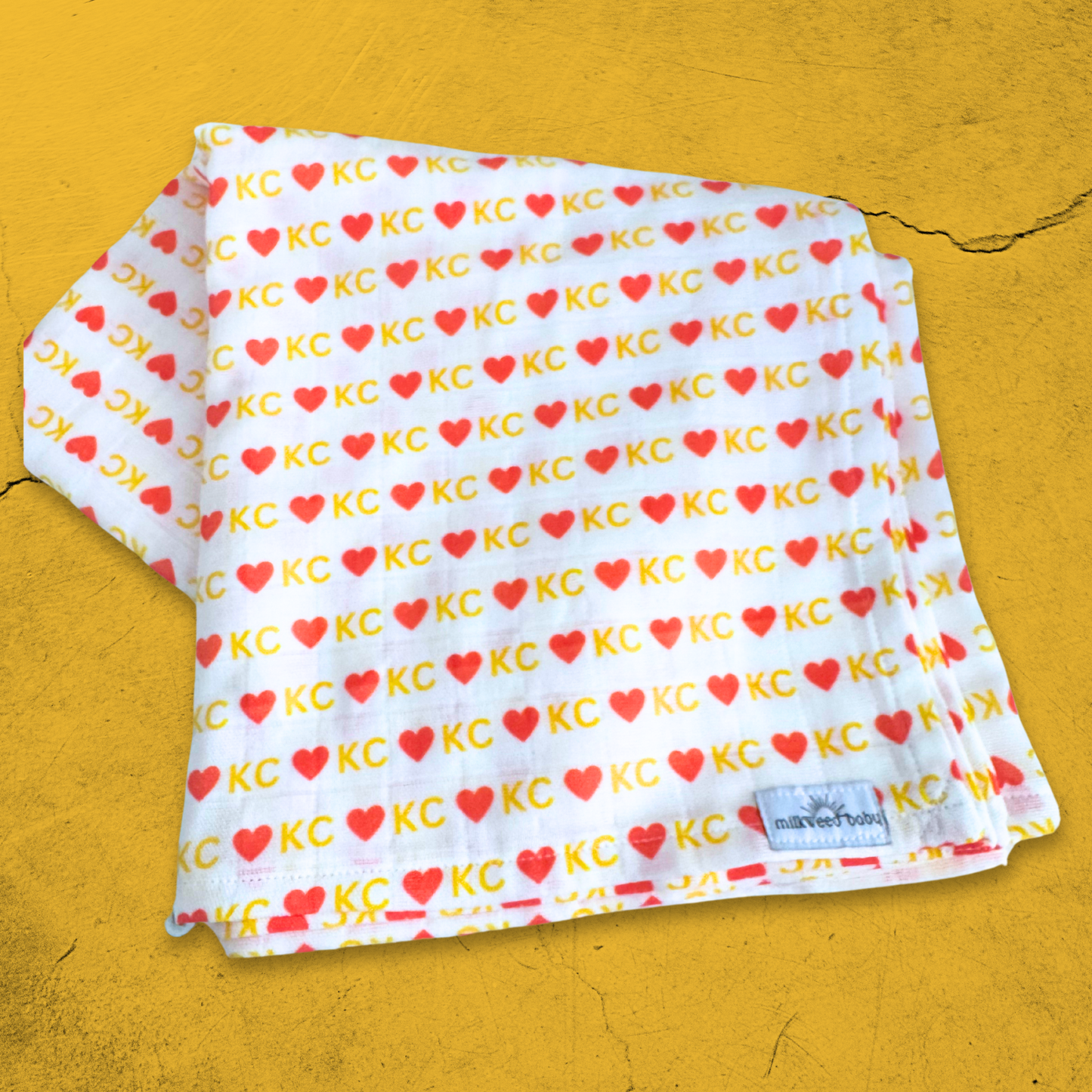 Folded KC Chiefs baby swaddle blanket, featuring a red and gold heart "KC" pattern on organic gauze fabric, displayed on a textured gold background.