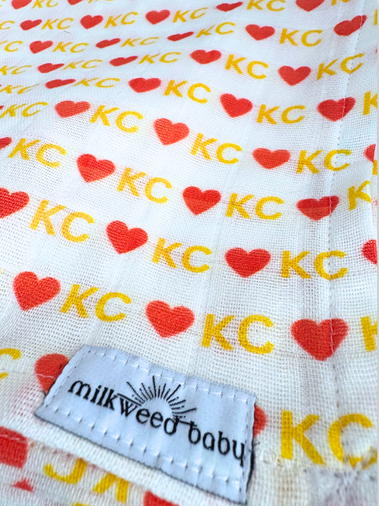 Close-up of a KC Chiefs-inspired baby swaddle featuring a repeating pattern of red hearts and "KC" in gold, printed on soft, organic Sweet Pea Gauze fabric.