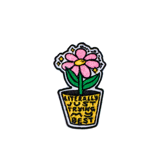 A close-up of an embroidered iron-on patch featuring a pink flower in a pot with the words "Literally Just Trying My Best" in black and yellow embroidery.
