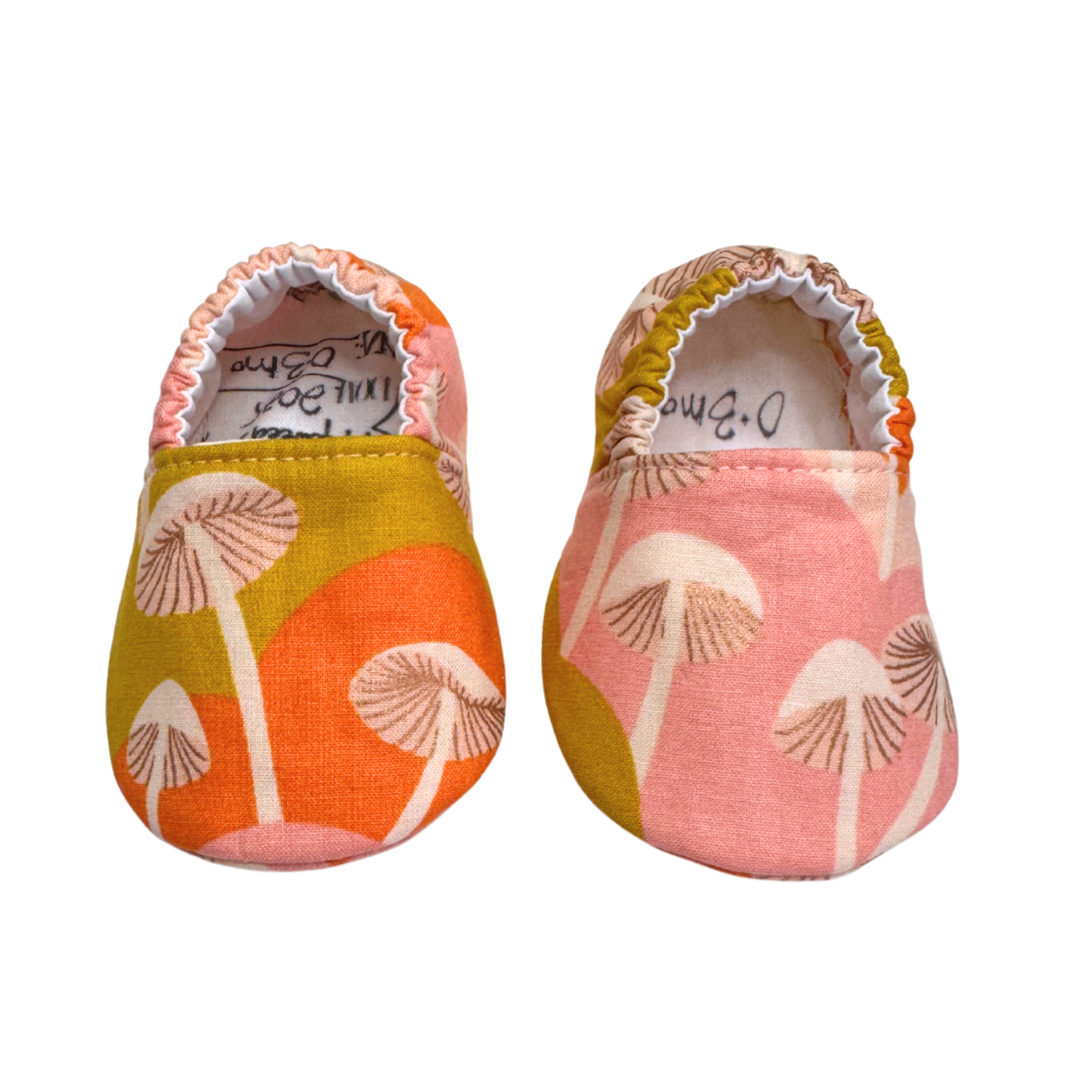 Handmade baby moccasins with a vibrant mushroom print in pink, orange, and mustard, featuring an elastic back and soft sole, size 0-3 months.