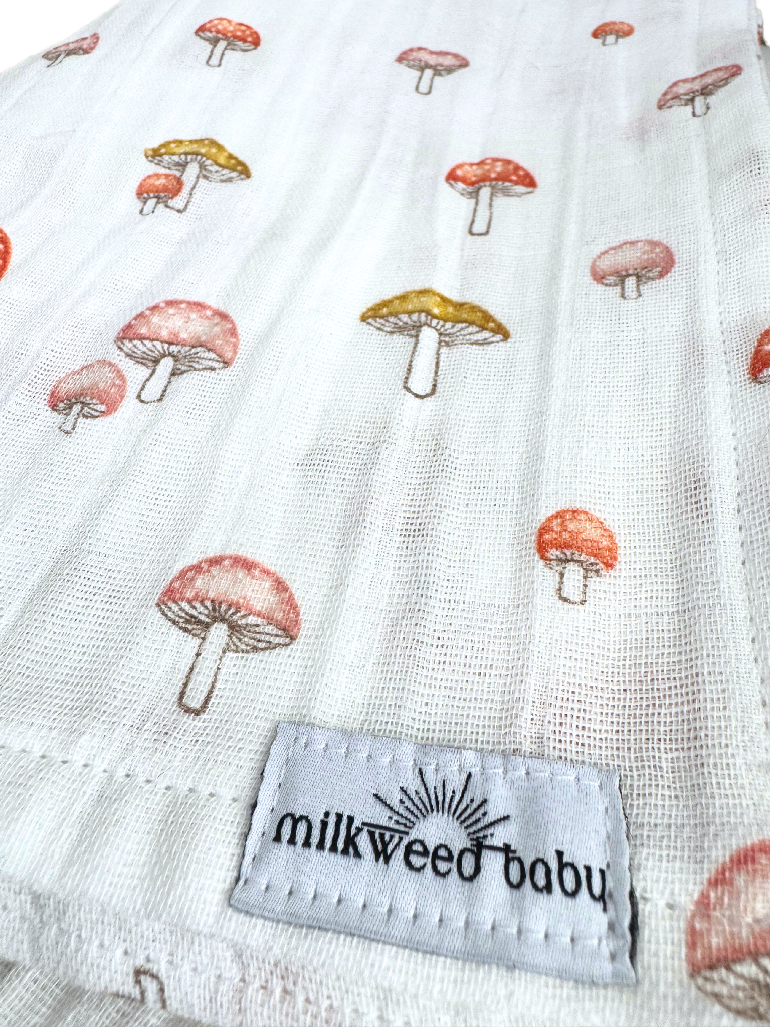 Close-up of a cotton gauze baby swaddle featuring a whimsical red and gold mushroom pattern, stitched with a "Milkweed Baby" tag.