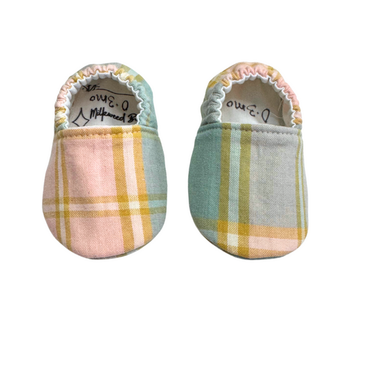 Handmade baby moccasins featuring a soft pastel plaid print in pink, blue, and golden yellow, with a soft sole and elastic back, size 0-3 months.