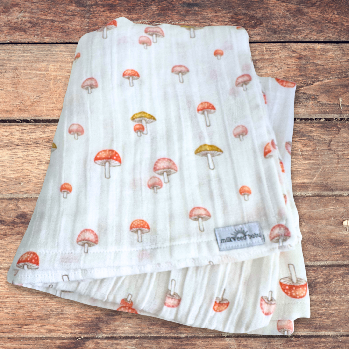 Folded mushroom-patterned baby swaddle made of soft cotton gauze, featuring red and gold mushrooms on a white background, displayed on a rustic wood surface.