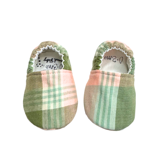 Handmade baby moccasins with a soft sage green and blush plaid print, featuring a soft sole and elastic back for a snug fit, size 0-3 months.