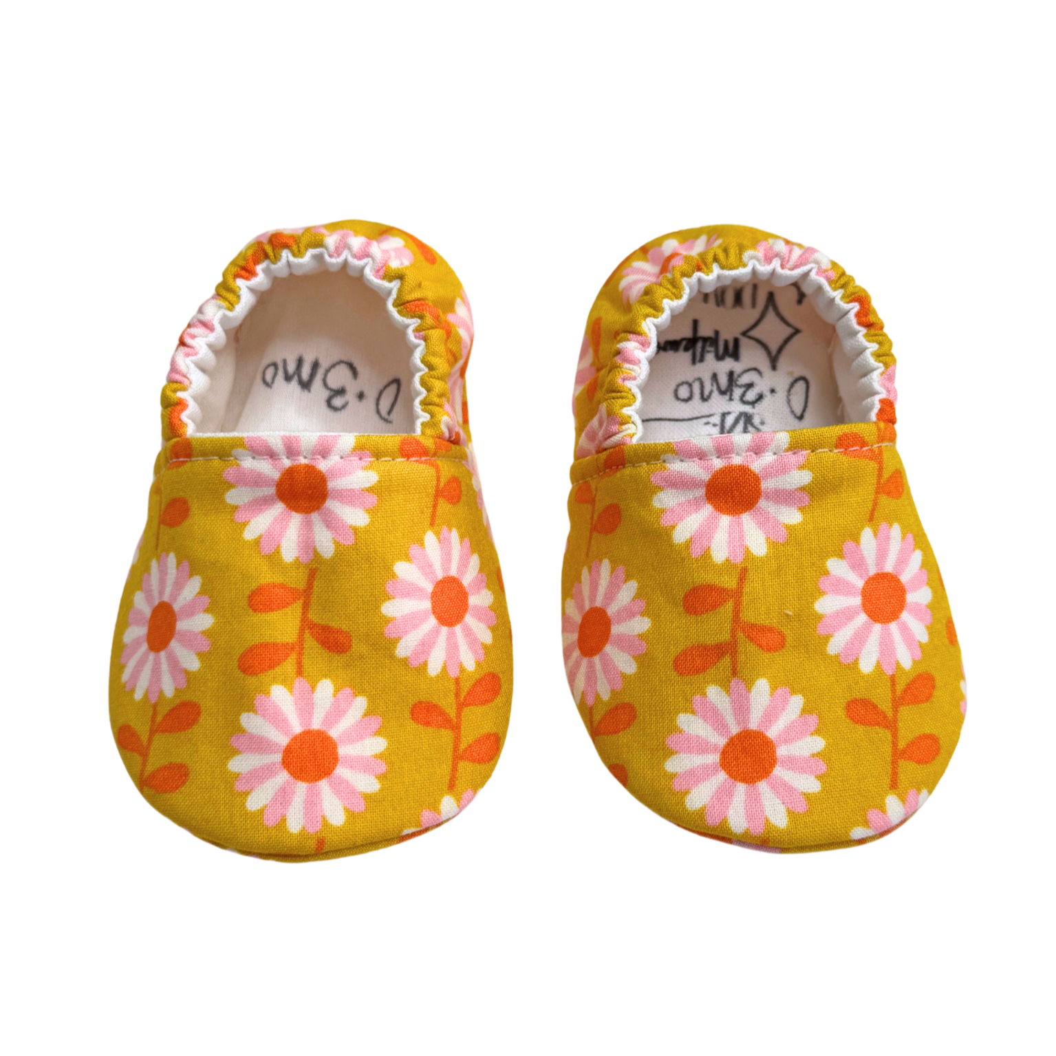 Handmade baby moccasins with a golden yellow daisy print, featuring pink and white flowers, a soft sole, and an elastic back for a snug fit, size 0-3 months.