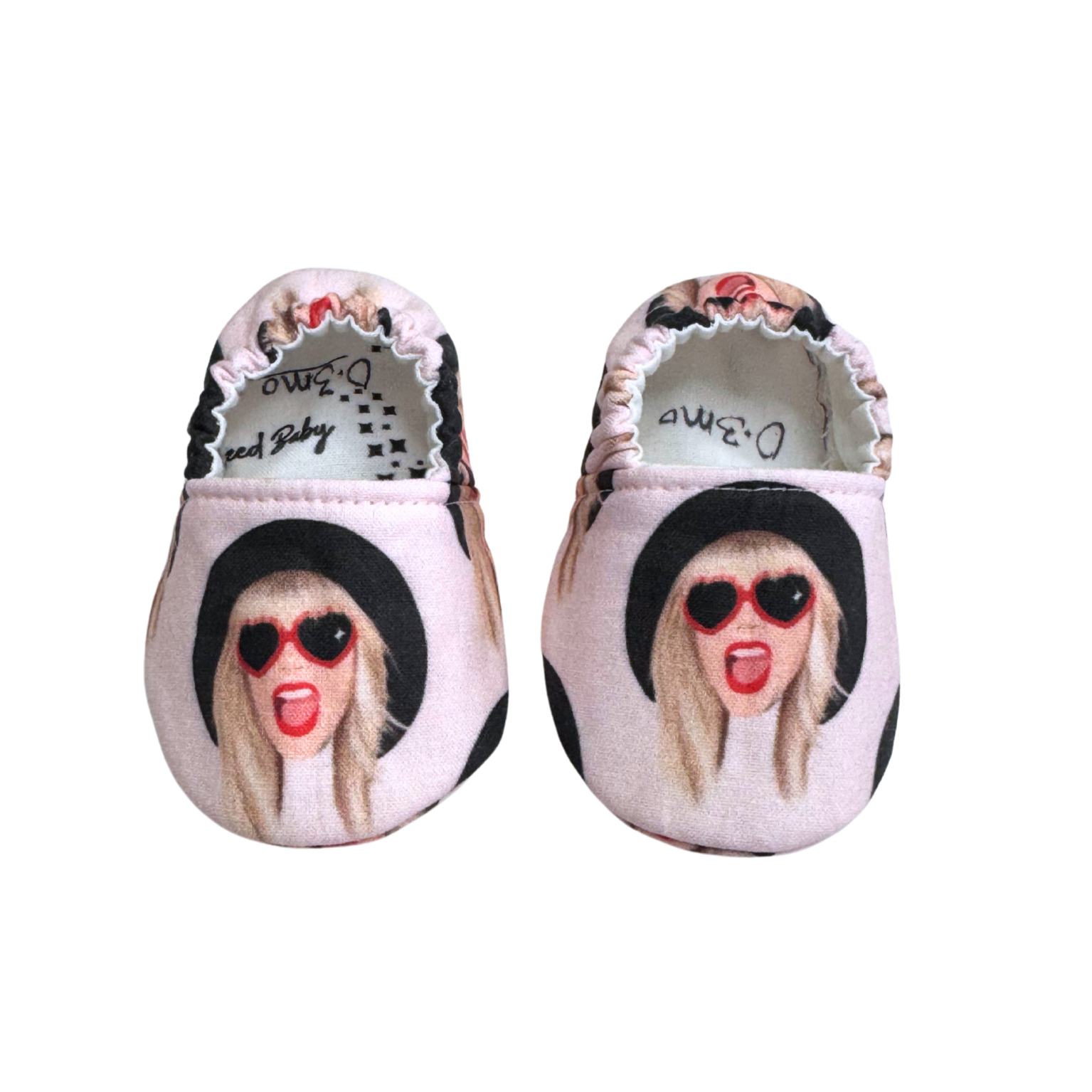 Handmade baby moccasins featuring a pink background with a Taylor Swift-inspired design, heart sunglasses, and red lipstick, soft sole, size 0-3 months.