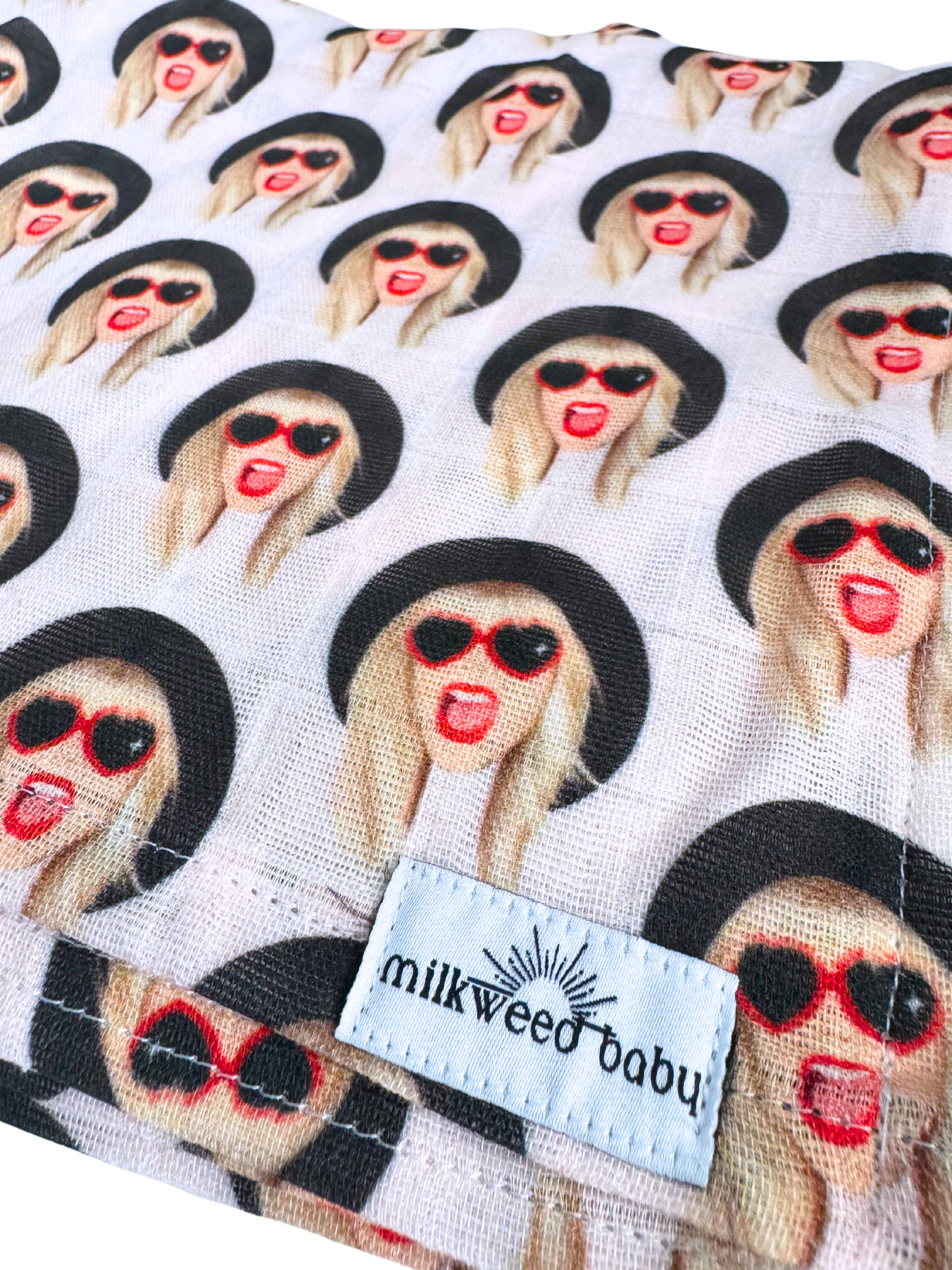 Close-up of a Taylor Swift-inspired baby swaddle blanket with a repeating print of a Taylor wearing red heart sunglasses and a black hat. The fabric is soft, breathable muslin gauze with a "Milkweed Baby" tag.
