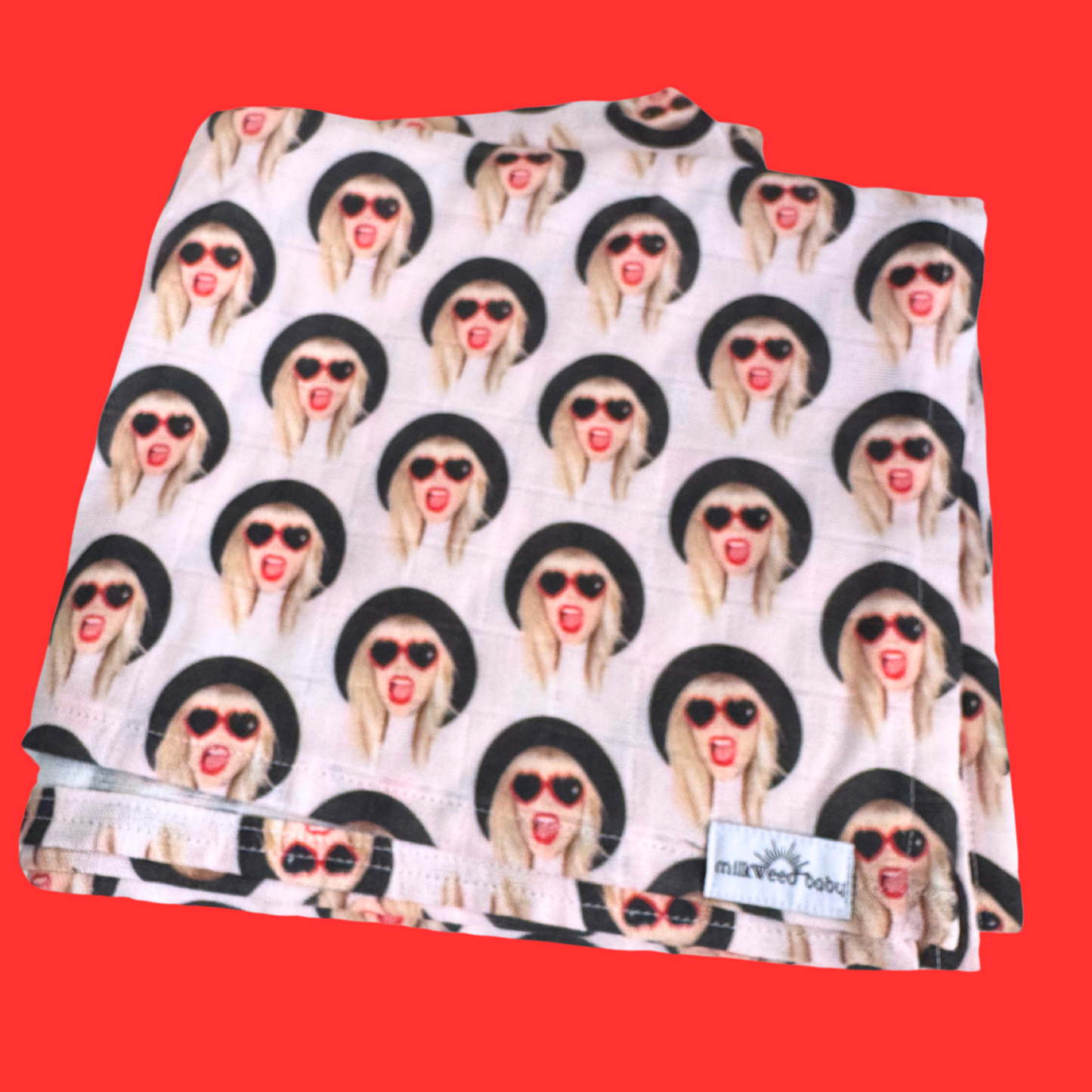 Taylor Swift-inspired baby swaddle blanket displayed against a red background, featuring a repeating design of Taylor with heart sunglasses and red lipstick. The swaddle is handmade and made of soft, crinkly gauze fabric.

